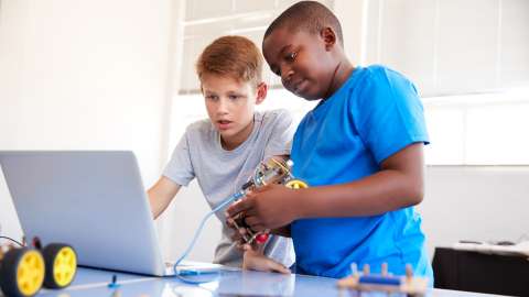 Two middle school students stand at a classroom worktable collaborating in front of a laptop they are using to program a connected Arduino device.